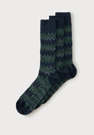 Drie paar donkerblauwe sokken met groene en witte zigzag- en gestreepte patronen, naast elkaar gerangschikt op een witte achtergrond.