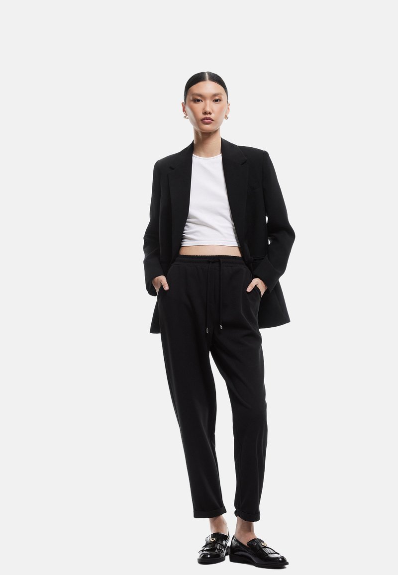 Sort skræddersyet blazer over en hvid cropped skjorte, parret med sorte snørespants og skinnende sorte sko. Minimalistisk design og glat tekstur.