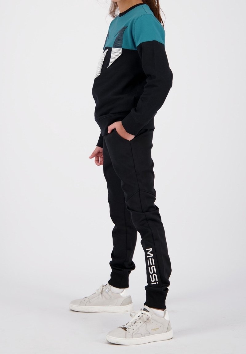Enfant portant un pantalon de jogging noir avec le texte "MESSI", un sweat-shirt noir et turquoise, des chaussettes blanches et des baskets blanches, debout les mains dans les poches.