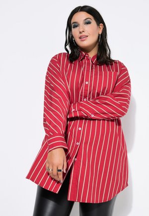 Kvinde med mørkt hår iført store øreringe, blå øjenskygge, oversized rød skjorte med hvide lodrette striber og sorte læderbukser, poserer selvsikkert.