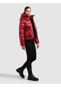 Nainen pukeutuneena kiiltävään punaiseen untuvatakkiin, mustiin legginsseihin ja mustiin polven korkeisiin saappaisiin, seisomassa yksinkertaisen vaalean taustan edessä.