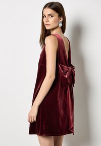 Burgundowa sukienka z weluru z głębokim dekoltem na plecach i dużą kokardą w pasie. Kształt w literze A i gładka tekstura.