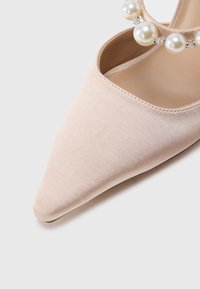 Γόβα με μυτερό πέλμα σε απαλό μπεζ σατέν με ομαλή υφή. Διαθέτει διακοσμητικό λουράκι διακοσμημένο με στρώσεις από μαργαριτάρια και διαυγείς χάντρες.