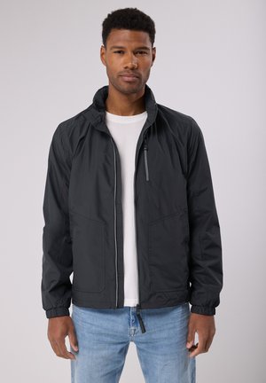 Zwarte lichtgewicht jas met een rits aan de voorkant, zijzakken, elastische manchetten en een opstaande kraag, gedragen over een wit shirt.
