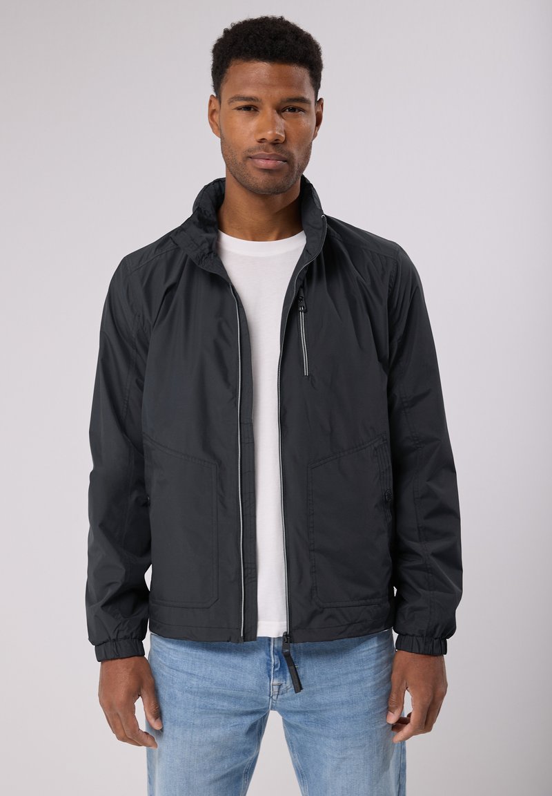 Schwarze leichte Jacke mit einem Reißverschluss vorne, Seitentaschen, elastischen Bündchen und einem Stehkragen, getragen über einem weißen Hemd.
