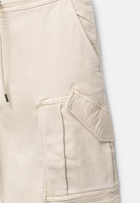 Pantaloni cargo beige realizzati in tessuto morbido, caratterizzati da numerose tasche, cuciture in vista e una vestibilità rilassata. La texture appare liscia.