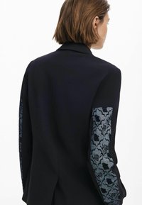 Black blazer featuring textured blue floral patterned fabric on sleeves, tailored fit, notched lapel, and a vent at the back.