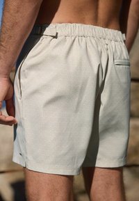 Patterned swim shorts with an elastic waistband, featuring a wavy design in light colors. Includes a small side pocket and smooth texture.