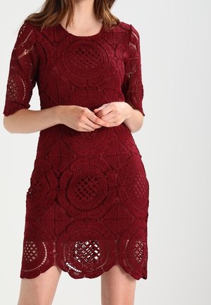 Frau trägt ein knielanges burgunderrotes Spitzenkleid mit kreisförmigen Mustern und halblangen Ärmeln, steht mit zusammengelegten Händen.