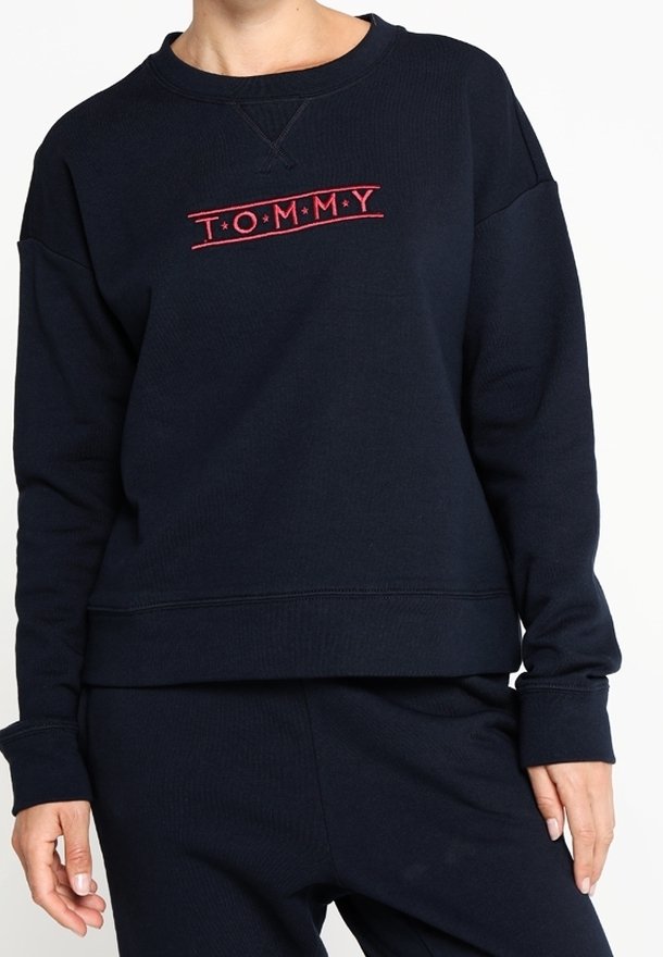 Personne portant un sweat-shirt bleu marine avec "TOMMY" en rouge, assorti d'un pantalon bleu marine, visible du cou jusqu'aux hanches.