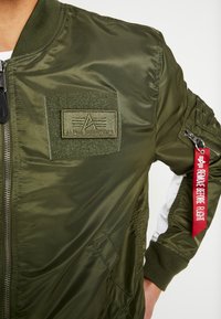 Olivgrüne Bomberjacke mit Reißverschluss, gerippten Bündchen und einem Patch auf der Brust. Mit einem roten "REMOVE BEFORE FLIGHT"-Tag.