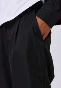 Pantalons noirs à finition lisse avec une coupe décontractée, dotés de poches latérales, de détails de plis et d'une taille visible au-dessus d'un haut blanc.