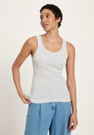 Frau trägt ein hellgraues ärmelloses Oberteil und lockere blaue Jeans, schaut seitlich und hält mit der Hand den Saum des Oberteils.