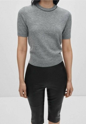 Henkilö, joka pukeutunut lyhythihaiseen harmaaseen neulepaitaan ja mustiin, vartaloa myötäileviin puolipitkiin housuihin, seisoo yksiväristä taustaa vasten.