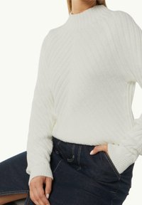 Un pull en tricot blanc avec un col haut, présentant une texture côtelée et des motifs diagonaux. Associé à une jupe en denim sombre au design minimal.