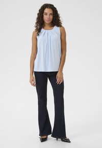 Gaiši zila bezpiešu topa ar krokotām detaļām, saskaņots ar tumšajiem platmalām džinsiem un melnām apaviem ar smailiem purngaliem. Gluda auduma, brīva piegriezuma.