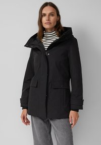 Vrouw in een zwart hoodie-jack over een gestreepte coltrui en grijze jeans, staand tegen een effen lichtgrijze achtergrond.