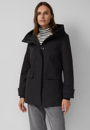 Vrouw in een zwart hoodie-jack over een gestreepte coltrui en grijze jeans, staand tegen een effen lichtgrijze achtergrond.