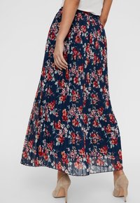 Kvinna i en marinblå plisserad midikjol med röd och vit blommig tryck och beige högklackade skor, stående med en hand på höften.