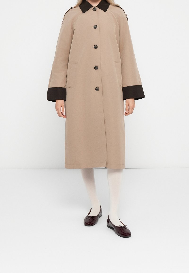 Femme portant un manteau long beige avec un col et des poignets noirs, des collants blancs et des ballerines rouge foncé, debout devant un fond blanc.