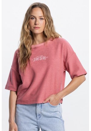 Blonde vrouw met lang golvend haar, gekleed in een losse roze top met korte mouwen en de tekst "JNSN", en een lichtblauwe spijkerbroek, hand in broekzak.