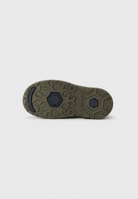 Suela de un zapato verde oliva con acentos hexagonales negros y un patrón de banda de rodadura geométrico sobre un fondo claro.