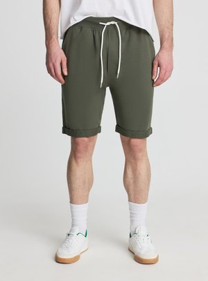 Mężczyzna w oliwkowo-zielonych szortach z wywiniętym dołem i białym sznurkiem, białych skarpetkach, białych trampkach i białej koszulce, stojący na jednolitym tle.