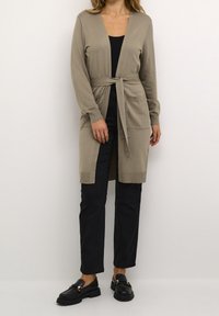 Donna che indossa un cardigan lungo color taupe chiuso in vita, una maglia nera, pantaloni neri e mocassini neri, in piedi contro uno sfondo neutro.