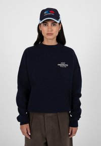 Jeune femme portant un pull bleu marine et une casquette assortie, tous deux étiquetés "PROHIBÉ", posant devant un fond gris clair uni.