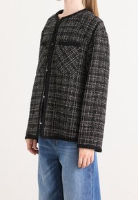 Femme portant une veste texturée à carreaux noir et blanc avec des poches avant sur un jean bleu sur fond blanc.