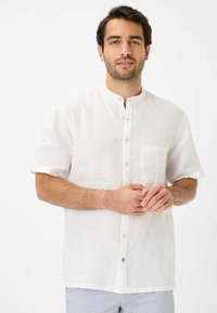 Camicia bianca di lino a maniche corte con colletto alla mandarino, chiusura con bottoni, taschino singolo sul petto e una trama sottile. Accento blu sui bottoni.