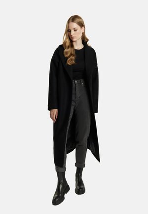 Vrouw met lang blond haar die een lange zwarte jas draagt, een zwart topje, donkerdere high-waisted jeans en zwarte chunky enkellaarzen.