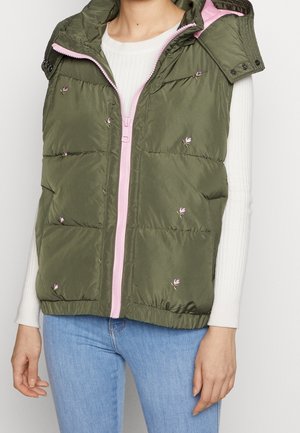 Oliwkowa pikowana kamizelka z delikatnym różowym haftem kwiatowym i jasnoróżowym zamkiem błyskawicznym, noszona na białym, prążkowanym, długim rękawie oraz niebieskich dżinsach.