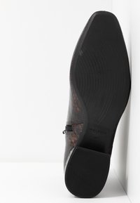 Semelle d'une bottine à bout pointu, de couleur brun foncé avec un motif en crocodile, talon bloc, présentée debout sur un fond blanc.