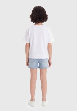 Barn med kort krøllet hår står med ryggen til, iført en hvid kortærmet skjorte, lyse denimshorts og hvide sneakers.