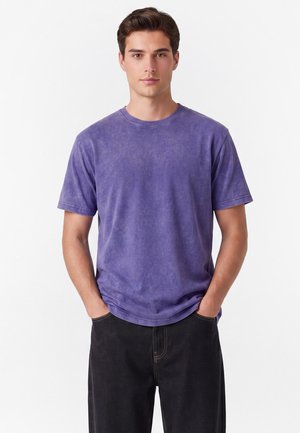 Nuori mies, jolla on lyhyet ruskeat hiukset, pukeutunut haalistuneeseen violettiin lyhythihaiseen t-paitaan ja mustiin housuihin, kädet taskuissa.