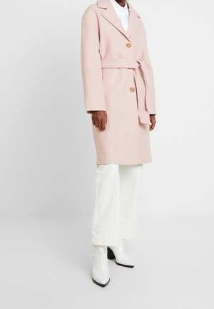 Henkilö, jolla on vaaleanpunainen vyöllinen takki, jossa on kaksi nappia, valkoiset housut ja valkoiset kärjestä terävät nilkkurit yksiväristä valkoista taustaa vasten.