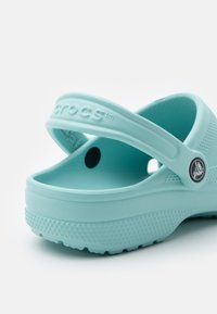 Crocs de cor azul claro com parte superior em plástico texturizado, tira traseira e sola salpicada, apresentando um detalhe de logótipo na lateral.