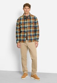 Karierte Flanelljacke in Braun, Blau und Grün; zwei Brusttaschen; schwarze Knöpfe; kombiniert mit beigen Hosen und braunen Turnschuhen.