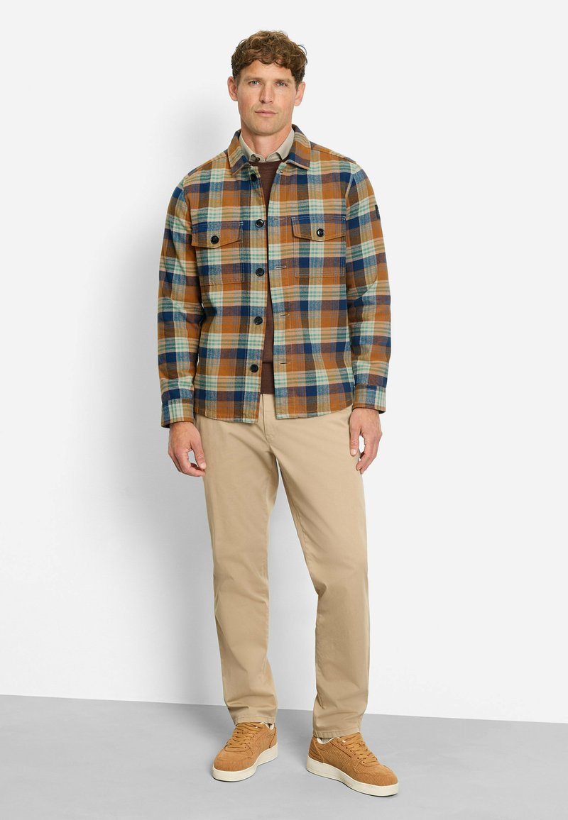 Karierte Flanelljacke in Braun, Blau und Grün; zwei Brusttaschen; schwarze Knöpfe; kombiniert mit beigen Hosen und braunen Turnschuhen.