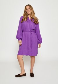 Lila Kleid mit strukturierter Oberfläche, langen Ballonärmeln und gebundenem Taillenband. Kombiniert mit dunkelbraunen flachen Schuhen.