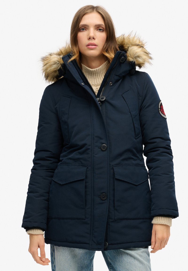 Parka aislante de color azul marino con capucha forrada de piel, cierre frontal con botones, dos grandes bolsillos y puños de punto acanalado para mayor calidez.