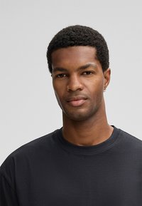 Junger Mann mit kurzem, lockigem Haar, der ein schwarzes Rundhals-T-Shirt und einen kleinen Stecker trägt und mit neutralem Ausdruck frontal blickt.