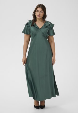 Mujer con un vestido largo de satén verde oscuro con mangas cortas con volantes y escote en V, combinando con zapatos de tacón negros puntiagudos, de pie.