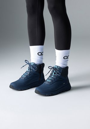 Personne portant des leggings noirs, des chaussettes blanches avec des symboles d'infini noirs, et des baskets montantes bleu marine avec des lacets bleus sur un sol gris.