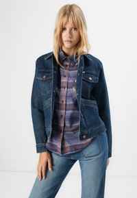 Ung kvinna som bär en mörk denimjacka över en lila rutig skjorta och ljusblå jeans, stående mot en vanlig vit bakgrund.