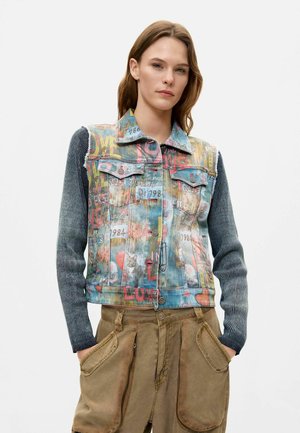 Giovane donna che indossa un gilet in denim multicolore con maniche lunghe scure a costine e pantaloni cargo marroni con grandi tasche.