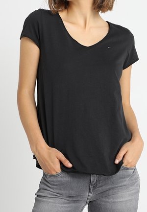 Frau trägt ein schlichtes schwarzes T-Shirt mit V-Ausschnitt und kurzen Ärmeln, die Hände in den Taschen, kombiniert mit ausgewaschenen grauen Jeans.