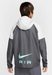 Nike vindjakke i grå og hvid med justerbar hætte, elastiske manchetter og stort blåt "AIR" logo på ryggen.
