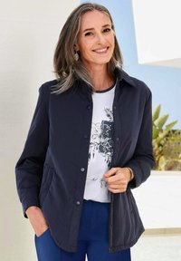 Navyblaues Hemdjacke, leichtes Material, mit zwei seitlichen Taschen, kombiniert mit einem weißen T-Shirt mit Blumenmuster und blauen Hosen.